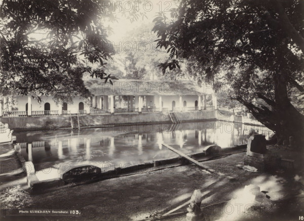 Bathing facility with gallery, possibly in Surabaya, photograph, Part of Travel Album with Forty Photos of the Dutch East Indies., number: '10', number: '105', photographer: Onnes Kurkdjian, Surabaya, 1912, Java, East Java, paper, daylight gelatin silver print, height 173 mm x width 236 mm, public baths, Dutch East Indies, The, A shaded scene of a long single-story building with a tiled roof and a covered veranda supported by columns, positioned along the far side of a calm rectangular pool that reflects the building and surrounding trees; a set of wooden steps leads from the veranda down to the water, and balustrades run along the veranda edge. Large overhanging tree branches frame the top of the image while a massive tree trunk and its exposed roots occupy the right foreground. In the near foreground a paved area with low stone barriers and worn steps slopes toward the pool, and a narrow plank lies across the edge of the water. The overall image shows dappled light and shadow across ...
