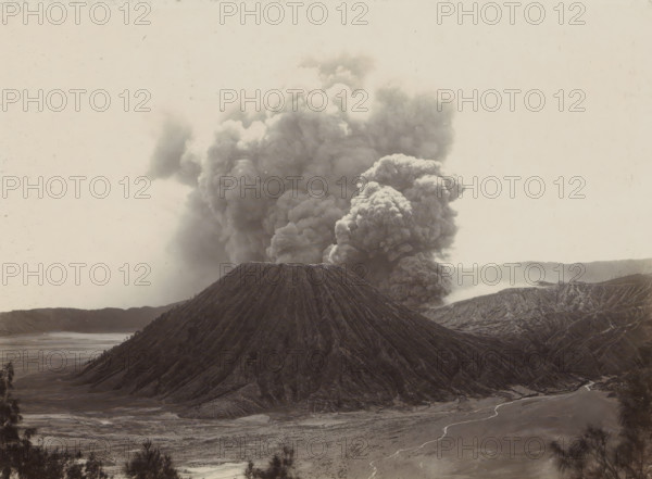 Smoking volcano in the Dutch East Indies, photograph, Part of Travel Album with Forty Photos of the Dutch East Indies., photographer: Onnes Kurkdjian, Dutch East Indies, The, 1912, paper, daylight gelatin silver print, height 175 mm x width 238 mm, volcano, A large conical mountain with steep, deeply ridged slopes sits in the foreground, its summit appearing flat and dark; behind and rising from its base is a massive dense plume of billowing smoke and ash that fills much of the sky with rounded, turbulent shapes; surrounding terrain shows eroded ridges and valleys with a wide flat plain in front, a winding pale stream crossing the plain, and distant low hills along the horizon; the overall palette is muted and monochromatic with textured surfaces and a hazy atmosphere.