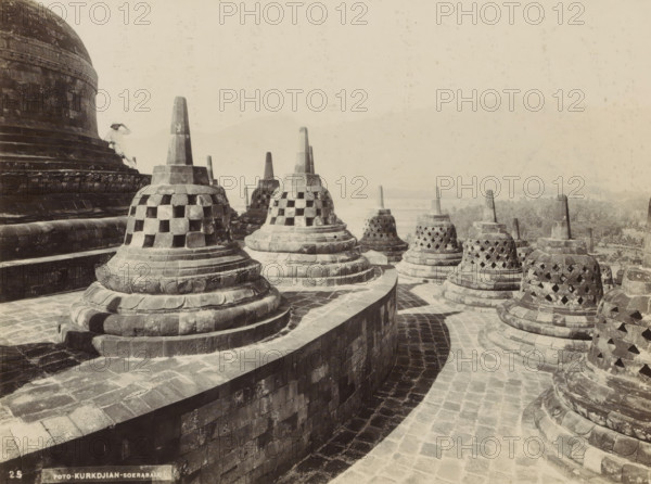 Stupas of Borobudur on Java, photograph, Part of Travel Album with Forty Photos of the Dutch East Indies., number: '25', photographer: Onnes Kurkdjian, Borobudur, 1912, Java, Central Java, paper, daylight gelatin silver print, height 178 mm x width 236 mm, temple, shrine, Hinduism, Buddhism, Jainism, Stone bell-shaped stupas with lattice openings sit atop tiered circular bases arranged along curved stone walkways, their pointed pinnacles rising above carved moldings and layered platforms. A large domed structure with carved bands and weathered blocks occupies the left side, and a person wearing light clothing and a broad-brimmed hat stands on its steps. The stone surfaces show varied tones and weathering, and distant vegetation and faint hills appear on the horizon under a pale sky.