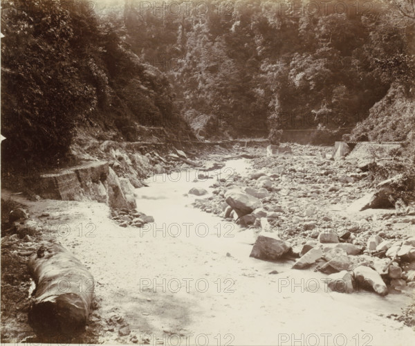 Stream bed with a road in the Dutch East Indies, album page, photograph, Part of Album with photos of railways in the Dutch East Indies and postcards., Dutch East Indies, The, c. 1880 - c. 1910, photographer, pasteboard, paper, albumen print, height 277 mm x width 232 mm, railway, train, brook, A wide rocky riverbed flanked by steep forested hills, with scattered boulders and stones along the shore and in the shallow water, a narrow path or ledge running along the left side beside exposed rock faces, a large fallen log resting near the foreground on the left, dense trees and foliage covering the slopes in the background, and a small man-made structure near the mid-right bank beside the rocky embankment.