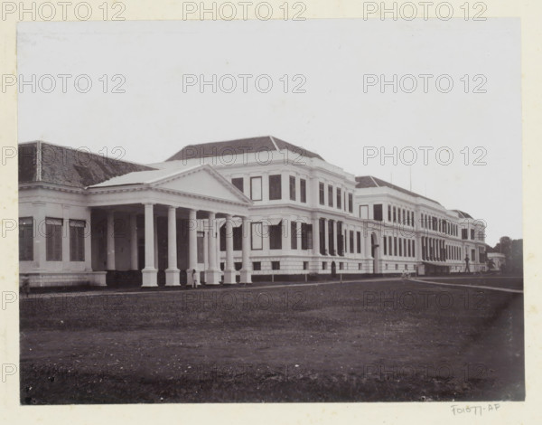 Waterlooplein Palace, Exterior of Daendels' palace on Waterlooplein in Batavia, album page, photograph, Part of Album with photos of Batavia and of the Ursuline sisters., Batavia, c. 1890 - c. 1910, photographer, Java, West Java, paper, pasteboard, daylight collodion silver print, height 170 mm x width 224 mm, palace, A large classical white building stretches across the image with a colonnaded portico at the left featuring tall round columns and a triangular pediment, adjoining a multi-story wing with rows of tall rectangular windows and decorative cornices; the building has a tiled hipped roof and several projecting bays, and stands behind a broad expanse of lawn in the foreground, with a few small indistinct figures near the portico and along the façade.