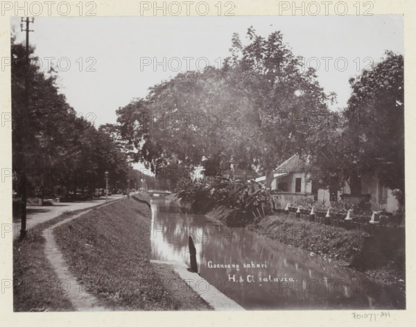 View of a house and the water of Gunung Sahari in Batavia, Goenoeng sahari., album page, photograph, Part of Album with photos of Batavia and of the Ursuline sisters., inscription: 'Goenoen sahari', photographer: Otto Hisgen, Batavia, c. 1890 - c. 1910, Java, West-Java, paper, pasteboard, daylight collodion silver print, height 165 mm x width 221 mm, dikes, levee, canals, waters (in city), A narrow waterway runs through the scene with still reflective water bordered by grassy banks and a dirt path on one side, tall leafy trees line both sides creating a canopy over parts of the water, a small bridge is visible in the distance, a modest building with a tiled roof and windows sits near the water under the trees, a row of short posts with cone-shaped tops lines the near bank in front of the building, dense foliage and plants grow along the water’s edge, a lamppost and scattered street elements appear along the path among the trees.