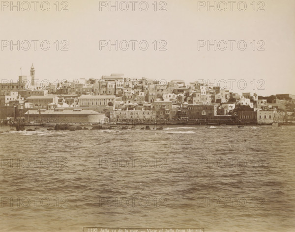 Jaffa, seen from the sea, Jaffa vu de la mer. - View of Jaffa from the sea., photograph, Part of Travel Album with Photos of Jerusalem and Other Biblical Places., negative number: '1193', photographer: attributed to Maison Bonfils, c. 1867 - c. 1895, Ottoman Empire, Attributed to, pasteboard, paper, albumen print, height 222 mm x width 282 mm, prospect of city, town panorama, silhouette of city, A wide waterfront view showing a dense cluster of multi-level stone and plaster buildings along a shoreline, with flat roofs, domes, arched windows and doorways, and a tall tower with a pointed top rising above the other structures, all rendered in a sepia tone; in front of the buildings a narrow quay with small boats and masts is visible, dark rocks protrude at the waterline, and the foreground is filled with rippling sea water under a pale sky.