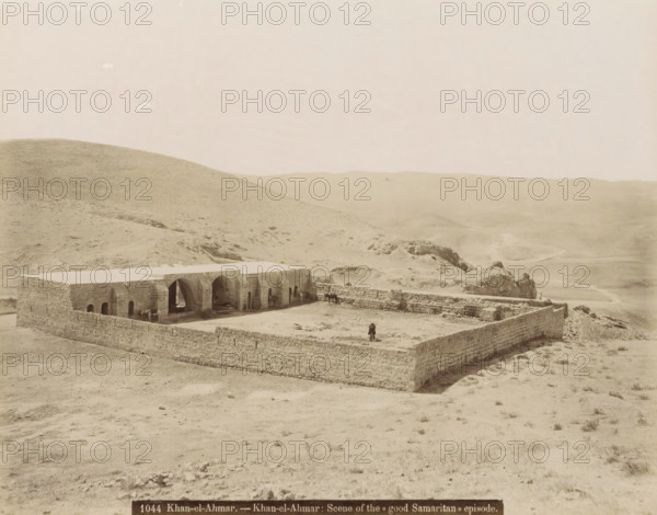 Khan al-Ahmar.- Khan al-Ahmar: Scene of the 'Good Samaritan' episode., Walled building in Khan al-Ahmar, photograph, Part of travel album with photos of Jerusalem and other biblical places., negative number: '1044', photographer: attributed to Maison Bonfils, Khan Al-Ahmar, c. 1867 - c. 1895, Palestine, West Bank, Attributed to, pasteboard, paper, albumen print, height 225 mm x width 283 mm, fence, wall, paling, desert, A stone enclosure sits in a barren landscape, its perimeter formed by low rough stone walls surrounding a rectangular courtyard and a long building with a flat roof and a series of arched openings along one side; inside the courtyard and near the arches are a few animals and figures, and a person stands near the outer wall; the surrounding terrain is dry and rolling with sparse vegetation, rocky outcrops, and distant undulating hills under a pale sky.