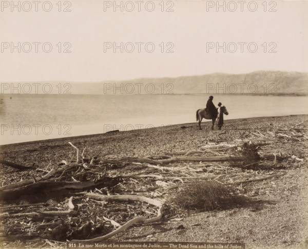 Rider and man on the shore of the Dead Sea with washed-up branches in the foreground, La mer Morte et les montagnes de Judée.- The Dead Sea and the hills of Judea., photograph, Part of Travel album with photos of Jerusalem and other biblical places., negative number: '911', photographer: Maison Bonfils, Dead Sea, c. 1867 - c. 1895, Jordan, pasteboard, paper, albumen print, height 227 mm x width 284 mm, beach, sea (landscape with figures, staffage), A wide shoreline scene with a pebbled beach strewn with pieces of driftwood in the foreground, a calm body of water extending to a distant mountain range under a pale sky, and a person mounted on a horse near the water's edge accompanied by another standing figure beside the horse.