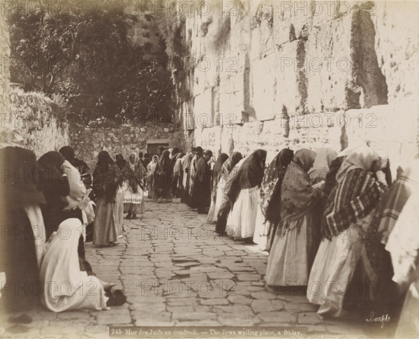 Believes praying at the Wailing Wall in Jerusalem, Mur des Juifs un vendredi. - The Jews waiting place, a Friday., photograph, Part of Travel album with photos of Jerusalem and other biblical places., negative number: '245', photographer: Maison Bonfils, Jerusalem, c. 1867 - c. 1895, pasteboard, paper, albumen print, height 0 mm x width 0 mm, prayer, praying, Jewish religion, Wailing Wall, A narrow stone-paved alley flanked on the right by a tall, light-colored stone wall and on the left by a lower rough stone wall and foliage, with a group of people standing or kneeling along the right wall and several others scattered along the left; many figures wear head coverings and layered garments with patterned shawls, some faces visible while others are turned toward the wall, and a few individuals stand in the center of the passage facing different directions.