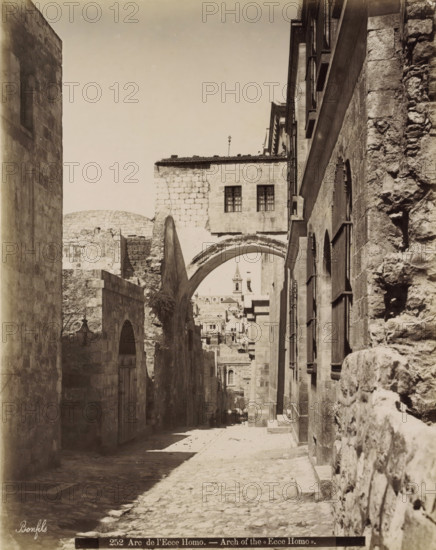 Arc de l'Ecce Homo. - Arch of the Ecce Homo., Ecce Homo arch in the Via Dolorosa in Jerusalem, photograph, Part of Travel album with photos of Jerusalem and other biblical places., negative number: '252', photographer: Maison Bonfils, Jerusalem, c. 1867 - c. 1895, Maison Bonfils, photographer, pasteboard, paper, albumen print, height 281 mm x width 226 mm, street, arch, archivolt, architecture, Via Dolorosa, A narrow cobblestone street flanked by tall stone buildings with arched doorways and barred windows, a large stone archway spanning the street with a rectangular stone structure above it that has two barred windows, a distant view through the arch of layered stone buildings and a tower with a pointed roof, lamp fixtures mounted on the walls, irregular stone textures on the facades, shadows falling across the street, and a photographer's signature visible near the lower edge.