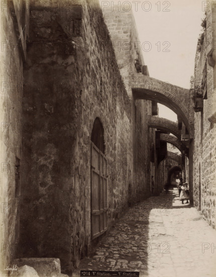 Fifth Station of the Cross on the Via Dolorosa in Jerusalem, V Station. - V Station., photograph, Part of Travel Album with Photos of Jerusalem and Other Biblical Places., negative number: '264', photographer: Maison Bonfils, Jerusalem, c. 1867 - c. 1895, pasteboard, paper, albumen print, height 282 mm x width 223 mm, street, stations of the cross, Via Dolorosa, A narrow cobblestone alley flanked by tall stone walls with multiple stone arches spanning overhead, wooden shutters and doors set into the walls, sunlight casting a bright strip along the center of the path while the sides remain in shadow, and several small figures farther down the alley including one adult and children wearing simple garments standing in the sunlit area.