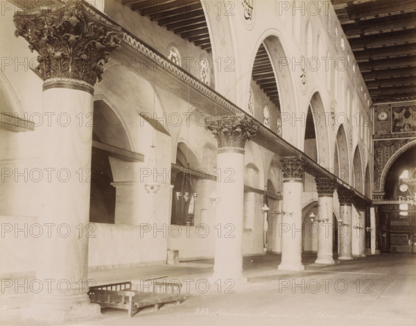 Jerusalem. Mosquée el-Aksa, - intérieur, Interior of the Al-Aqsa Mosque on the Temple Mount in Jerusalem, photograph, Part of Travel album with photos of Jerusalem and other biblical places., inscription: 'BFK Rives', negative number: '283', photographer: Maison Bonfils, Jerusalem, c. 1867 - c. 1895, pasteboard, paper, albumen print, height 225 mm x width 282 mm, temple, shrine, Islam, Mohammedanism, Al-Aqsa Mosque, A long interior view showing a row of tall cylindrical columns with ornate carved capitals supporting a decorated horizontal beam and a high ceiling with exposed wooden beams, a series of arched openings along the wall each topped by small circular windows, hanging pendant lights suspended from the ceiling at various heights, decorative painted or carved roundels on the arches, patterned detailing along the beam and arch faces, a wooden bench resting near the base of one column, and a richly decorated archway and panel work visible toward the far end.