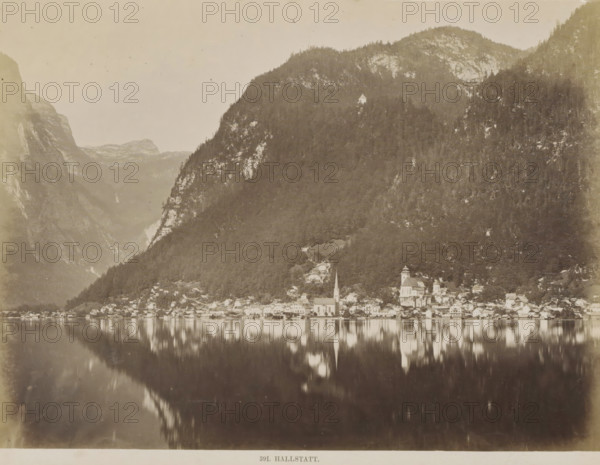 Hallstatt, View of Hallstatt with lake and surrounding mountains, photograph, Part of Travel album with photos of sights in Germany and Austria and of works of art., negative number: '391', c. 1870 - c. 1900, photographer, Upper Austria, paper, albumen print, height 198 mm x width 258 mm, prospect of village, silhouette of village, A wide lakeside scene showing a village of clustered buildings along a shoreline at the base of a steep, tree-covered mountain, with two church structures rising above the houses and their reflections visible in the calm water, the mountain slope dominated by dense forest and patches of exposed rock, a smaller building perched higher on the hillside, distant mountain ridges visible beyond and the entire scene rendered in a high-contrast, monochrome photographic style.