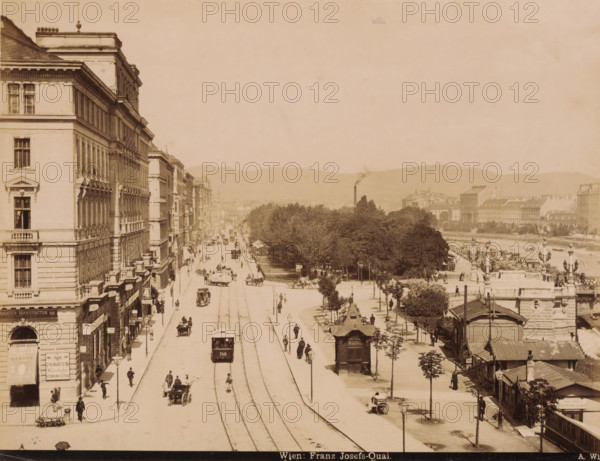 View of the Franz Josefs-Quai. in Vienna with shops, buildings, trams and carts, Wien. Franz Josefs-Quai., photograph, Part of Travel album with photos of sights in Germany and Austria and of works of art., photographer: A. Wimmer, Vienna, c. 1880 - c. 1895, Lower Austria, paper, albumen print, height 202 mm x width 264 mm, shop, store, tramway, tram, pier, quay, wharf, four-wheeled, animal-drawn vehicle, eg: cab, carriage, coach, street (city(-scape) with figures, staffage), A wide street scene showing a long avenue flanked by tall multi-story buildings on the left and a tree-lined promenade and riverbank on the right, with tram tracks running down the center of the street and a tram car moving along them; horse-drawn carriages and pedestrians are visible on the road and sidewalks, lamp posts line the avenue, and small kiosks and structures dot the promenade; in the distance low hills form the horizon and a smokestack rises above clustered buildings, while decorative facades, balconie...
