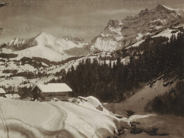 Landscape near Adelboden., View of the snow-covered mountains near Adelboden, photograph, Part of Travel album with photos of Germany, Switzerland, the Suez Canal and the Dutch East Indies., negative number: '469', photographer: Nikles, Adelboden, c. 1880 - c. 1900, paper, gelatin silver print, developed gelatin silver print, height 204 mm x width 261 mm, mountain-top, peak (landscape with figures, staffage), A wide snowy mountain scene with steep, jagged peaks in the background and forested slopes below, a few wooden cabins with snow-covered roofs near the lower left, a winding snow track in the foreground leading toward the cabins, dense dark evergreen trees covering the mid and right slopes, a partially visible stream bordered by snow and rounded rocks near the lower right, and scattered small outbuildings and clearings dotting the snow-covered valley.