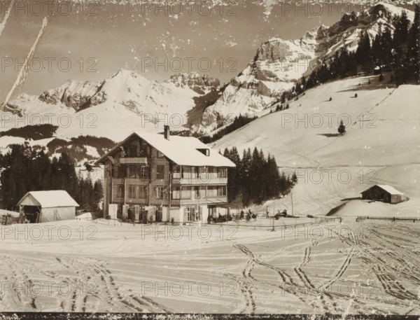 View of a chalet in the snowy mountains of Gilbach (Adelboden), Adelboden, Gilbach, photograph, Part of Travel album with photos of Germany, Switzerland, the Suez Canal and the Dutch East Indies., photographer: Nikles, c. 1880 - c. 1900, paper, gelatin silver print, developed gelatin silver print, height 198 mm x width 163 mm, rural housing, eg country-house, villa, cottage, mountain-top, peak (landscape with figures, staffage), A large multi-story wooden building with a snow-covered roof sits in front of a snowy landscape, its balconies and windows visible and a chimney rising from the roof; a smaller barn-like structure with a pitched roof stands nearby; tracks and paths crisscross the snow in the foreground; a line of dark evergreen trees frames the midground; steep snow-covered mountain slopes and rocky peaks rise behind the trees, with scattered lone trees on the slopes and patches of exposed rock.