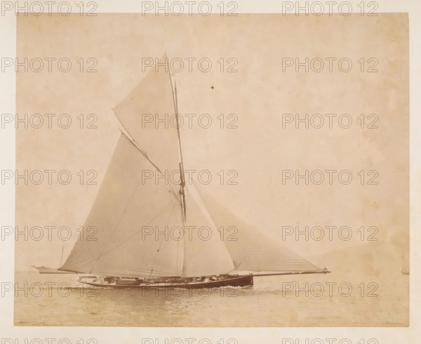 Sailing ship launched, album page, photograph, This photo is part of an album., check stamp: 'Adamson, Rothesay.H.B', photographer: James Adamson, c. 1880 - c. 1900, paper, albumen print, height 215 mm x width 272 mm, sailing-ship, sailing-boat, landscapes with waters, waterscapes, seascapes (in the temperate zone), A large sailboat shown in profile on calm water with tall sails raised and visible rigging, a long mast rising toward the top of the image, a sleek dark hull with details of the deck and small structures, thin lines of the sails showing seams and shading, a small boat alongside the main vessel, a faint distant shoreline on the left, and a pale, textured sky occupying much of the background.
