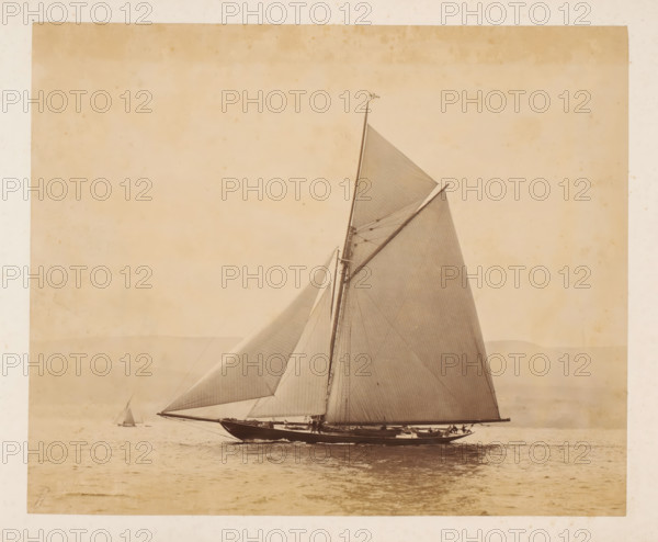 Two sailing ships launched, album page, photograph, This photo is part of an album., check stamp: 'Adamson, Rothesay.H.B', photographer: James Adamson, c. 1880 - c. 1900, paper, albumen print, height 229 mm x width 279 mm, sailing-ship, sailing-boat, landscapes with waters, waterscapes, seascapes (in the temperate zone), A large sailboat viewed in profile on calm water with its tall mast and multiple taut sails filling the right side of the image, a small flag at the mast top, rigging lines and spars visible, crew members positioned along the deck, a smaller distant sailboat on the left horizon, soft tonal gradations in the sky and faint landforms near the horizon, and the photograph framed by a light border with subtle aging marks.
