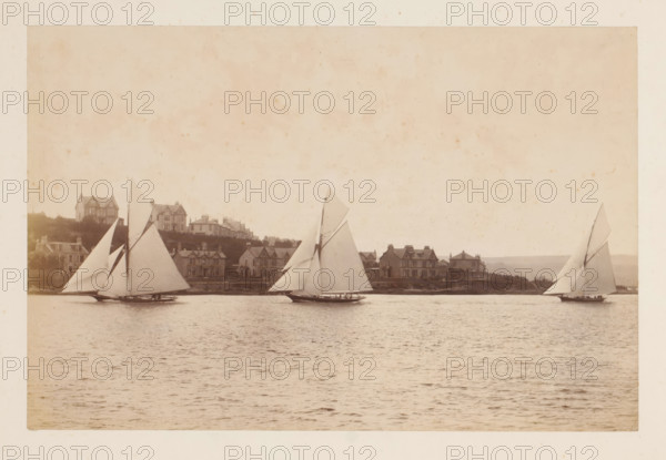 Sailing ships Yvonne, Doris and Darthula in the water with buildings in the background, album page, photograph, This photo is part of an album., check stamp: 'Adamson, Rothesay.H.B', photographer: James Adamson, c. 1880 - c. 1900, paper, albumen print, albumendruk, height 182 mm x width 284 mm, sailing-ship, sailing-boat, landscapes with waters, waterscapes, seascapes (in the temperate zone), civic architecture, edifices, dwellings, A sepia-toned scene of a calm body of water with three sailboats in the foreground, each boat showing tall triangular sails and low hulls with small groups of people aboard; along the distant shoreline a row of houses and larger buildings sit on a gentle rise with varied rooflines and chimneys, and grassy areas and trees are visible behind and between the structures; the sky above is light and mottled with faint spots and a textured appearance.