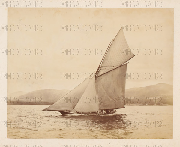 Sailing ship Idalia No. 1 launched with a hilly landscape in the background, album page, photograph, This photo is part of an album., check stamp: 'Adamson, Rothesay.H.B', photographer: attributed to James Adamson, c. 1880 - c. 1900, Attributed to, paper, albumen print, height 218 mm x width 279 mm, sailing-ship, sailing-boat, landscapes with waters, waterscapes, seascapes (in the temperate zone), (high) hill, A large sailboat pictured on a body of water with tall, triangular sails filled by wind, rigging lines visible against a light sky, a low shoreline and hills in the background, rippled water in the foreground, and a narrow strip of boat hull and crew partially visible near the waterline.