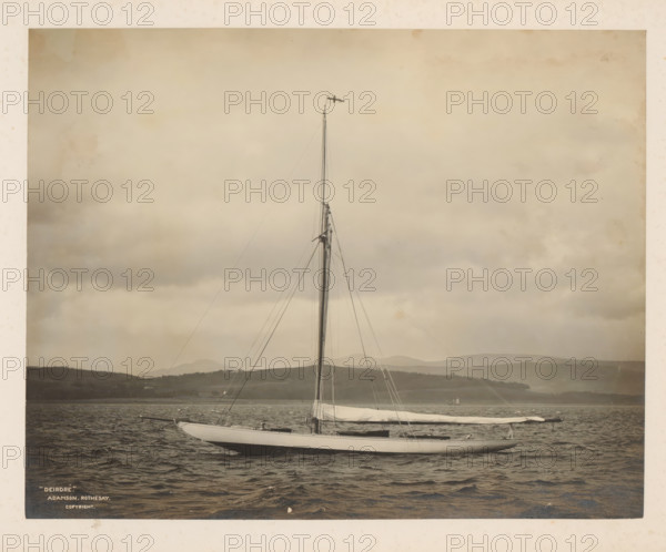 Sailing ship Deirdré in the water with a hilly landscape in the background, album page, photograph, This photo is part of an album., inscription: ''Deridré', Adamson. Rothesay., Copyright', check stamp: 'Adamson, Rothesay.HB, photographer: attributed to James Adamson, c. 1880 - c. 1900, James Adamson, photographer, Attributed to, paper, gelatin silver print, developing gelatin silver print, height 218 mm x width 279 mm, sailing-ship, sailing-boat, landscapes with waters, waterscapes, seascapes (in the temperate zone), (high) hill, A sailboat with a tall mast and multiple rigging lines is floating on rippled water, its sails furled and covered, with a small flag at the mast top; low rolling hills form the distant shoreline under a cloudy sky, and another small vessel is faintly visible near the horizon.