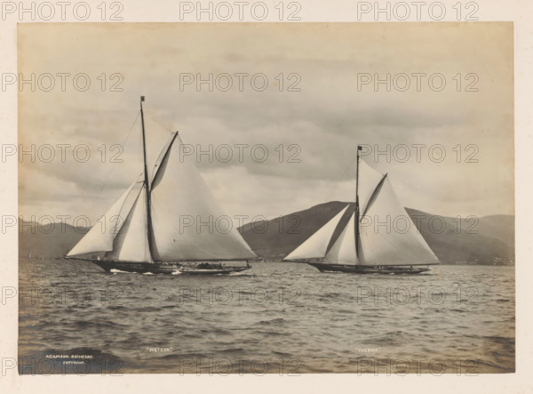 Sailing ships Meteor and Iverna at sea with a hilly landscape in the background, album page, photograph, This photo is part of an album., inscription: ''Meteor.', 'Iverna.'', inscription: 'Adamson, Rothesay, copyright', photographer: James Adamson, c. 1880 - c. 1900, paper, gelatin silver print, developed gelatin silver print, height 206 mm x width 290 mm, sailing-ship, sailing-boat, landscapes with waters, waterscapes, seascapes (in the temperate zone), (high) hill, Two large sailing yachts with tall masts and full sails are shown side by side on a calm sea, each hull dark against lighter water, with rigging lines visible along the masts and sails. The sails are pale and taut, showing fabric texture and subtle shading, and small groups of people appear along the decks. Low, rounded hills form a distant shoreline under a cloud-filled sky, and the surface of the water in the foreground shows gentle ripples and reflections.