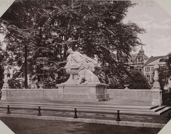 War monument in Hofgaren Park in Düsseldorf, album page, photomechanical print, This print is part of an album., inscription: 'Stengel & Co, 1897, Dresden', Düsseldorf, cliché maker: Stengel & Co., Dresden, photographer, North Rhine-Westphalia, Saxony, paper, collotype, photogravure, height 258 mm x width 355 mm, monument, statue, A stone monument sits on a raised rectangular platform with decorative railing posts in front; atop the platform a sculpted figure reclines on an ornate pedestal while a large carved lion rests at the base of the pedestal; the monument is flanked by urns and decorative posts on the platform edges; behind the monument a dense canopy of trees fills most of the background on one side while a multi-storied building with a domed roof and architectural detailing appears to the side; the foreground shows patterned paving and a low curb before a darker ground surface.