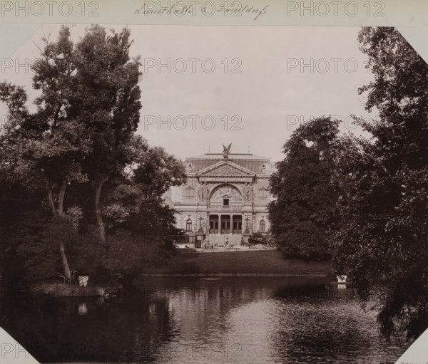 View of the Kunsthalle in Düsseldorf from 1891, album page, photomechanical print, This print is part of an album., annotation: 'Kunsthalle in Düsseldorf', inscription: 'Stengel & Co, 1897, Dresden', Düsseldorf, cliché maker: Stengel & Co., Dresden, photographer, North Rhine-Westphalia, Saxony, paper, collotype, photogravure, height 258 mm x width 355 mm, permanent exhibition, museum, A large neoclassical building with ornate carvings and a central pediment sits beyond a grassy bank, framed by tall leafy trees on both sides and reflected in a calm body of water in the foreground; a stone staircase leads up to the building's entrance where a few small human figures stand near doorways, and an angelic statue crowns the roof above sculptural reliefs, while dense foliage and tree trunks fill the image margins and the water shows rippled reflections of the scene.