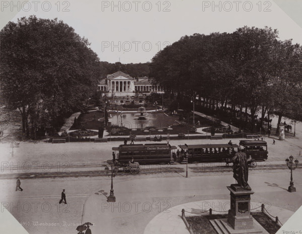 View of the Kurhaus in Wiesbaden, album page, photomechanical print, This print is part of an album., inscription: '4950. Wiesbaden, Kursaalplatz.', 1890 - 1899, paper, collotype, photogravure, height 258 mm x width 355 mm, public gardens, park (city(-scape) with figures, staffage), civic architecture, edifices, dwellings, Hessen, Wiesbaden, A wide plaza scene with a large neoclassical building centered in the background, flanked by rows of tall trees and a formal garden with paths, ornamental flowerbeds, and a rectangular reflecting pool with a central fountain. In front of the garden there are benches, lampposts, and a low decorative fence, while tram cars and a horse-drawn cart are positioned along the street in front of the plaza. Several pedestrians walk across the open paved area, and a large statue on a pedestal stands in the foreground with chained posts around its base. The image is monochrome and shows a broad view from an elevated vantage point.