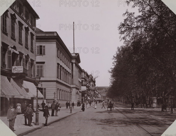 View of the Wilhelmstrasse in Wiesbaden, album page, photomechanical print, This print is part of an album., inscription: '3817. Wiesbaden, Wilhelmstrasse', 1890 - 1899, paper, collotype, photogravure, height 258 mm x width 355 mm, street (city(-scape) with figures, staffage), Hessen, Wiesbaden, A wide street scene with a row of multi-story buildings on the left featuring shuttered windows, awnings, a hanging shop sign, and decorative cornices, and a line of tall leafy trees on the right forming a shaded avenue; people in period clothing walk and stand along the sidewalks and street, some near lampposts and storefronts, while a cart with a horse is positioned on the roadway and tracks run down the center; the sky is clear above and a tall flagpole rises from one of the buildings.