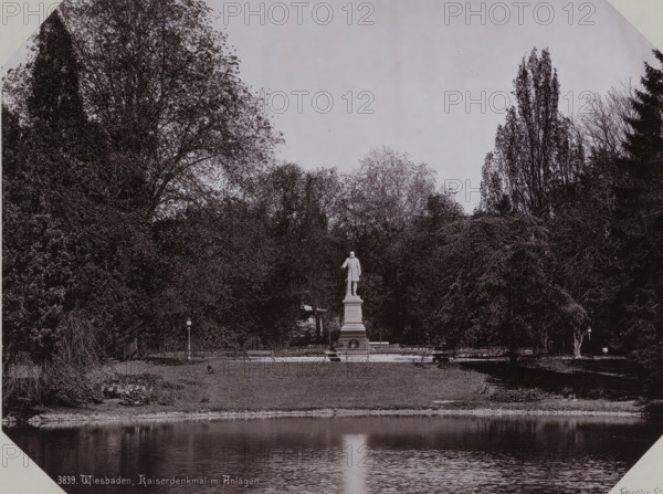 View of Warmer Damm city park with a statue of Emperor Wilhelm I in Wiesbaden, album page, photomechanical print, This print is part of an album., inscription: '3839. Wiesbaden, Kiserdenkmal m. Anlagen', 1890 - 1899, paper, collotype, photogravure, height 258 mm x width 355 mm, Person, Wilhelm I (Emperor of Germany), sculpture, public gardens, park (city(-scape) with figures, staffage), Hessen, Wiesbaden, A black and white scene of a park with a reflecting pond in the foreground, a grassy bank behind it, and a raised paved area with benches and light posts; at the center of the paved area stands a tall statue of a person on a multi-tiered pedestal; dense trees and tall foliage fill the background on both sides, framing the statue, and the water shows a vertical reflected highlight leading toward the statue.