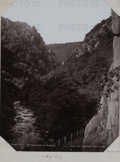View of the Bodethal in Thale, album page, photomechanical print, This print is part of an album., inscription: '3595. Thale, Blick . Kronentempel ins Bodethal., Verlag v. Zedler u. Vogel, Kunstanstalt Darmstadt 1898', publisher: Zedler und Vogel, Darmstadt, 1898, Hessen, paper, collotype, photogravure, height 258 mm x width 355 mm, ravine, chasm, abyss, canyon, Harz, A black and white photograph showing a narrow river flowing through a densely wooded valley flanked by steep rocky cliffs, with tree-covered slopes rising on both sides and jagged rock outcrops visible along the ridgelines, a rocky cliff face occupying the right foreground with a railing running along a path at its base, and an expanse of sky above the valley.