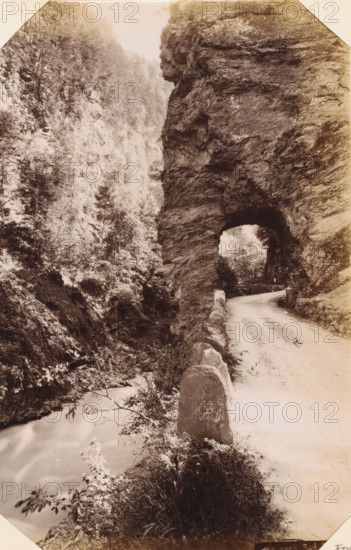 Taminakloof, album page, photograph, This photo is part of an album., annotation: 'Ragatz - Tamina Schlucht', c. 1885 - in or before 1892-04-29, photographer, paper, albumen print, height 209 mm x width 132 mm, ravine, chasm, abyss, canyon, A narrow dirt road curves into a tunnel carved through a tall rocky cliff, with a low stone barrier lining the road's outer edge; the tunnel opening reveals light and trees beyond while the cliff face shows layered rough textures and overhangs; opposite the road a steep forested slope rises with dense trees and undergrowth, and a stream flows at the base past rocks and vegetation; the scene is rendered in a sepia-toned photograph with soft contrasts and a vignette around the edges.