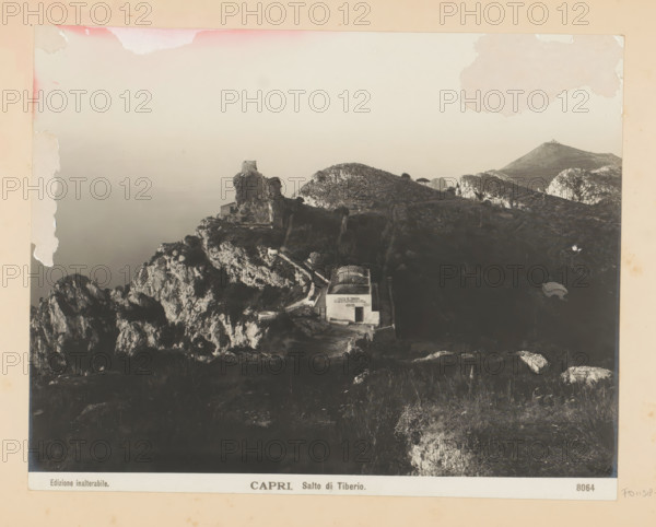 Capri. Salto di Tiberio, View of the Salto di Tiberio in Capri, photograph, Part of Photo album with recordings of sights and works of art in Italy., number: '8064', Capri, c. 1875 - in or before 1907, photographer, paper, gelatin silver print, developing gelatin silver print, height 190 mm x width 242 mm, coastal cliff or crag, Salto di Tiberio, A faded sepia photograph showing a rocky coastal landscape with cliffs and rugged hills descending toward a calm sea, a ruined stone structure perched atop a prominent rock outcrop, a small white building with an arched roof set into the hillside near a winding path, scattered vegetation on the slopes, distant rounded hilltops and rocky formations on the horizon, and areas of discoloration and peeling along the photograph's top and left margins.
