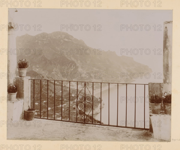 View of a bay (presumably) in Italy, photograph, Part of Photo album with photographs of sights and works of art in Italy., Italy, c. 1875 - in or before 1907, photographer, paper, daylight gelatin silver print, height 195 mm x width 243 mm, gulf, bay, A stone terrace with a metal railing featuring a central decorative scrollwork panel overlooks a coastal cliffside with terraced slopes and a distant shoreline; potted plants sit on short stone pillars at each side of the railing and on the terrace floor, and two tall, weathered columns frame the view.
