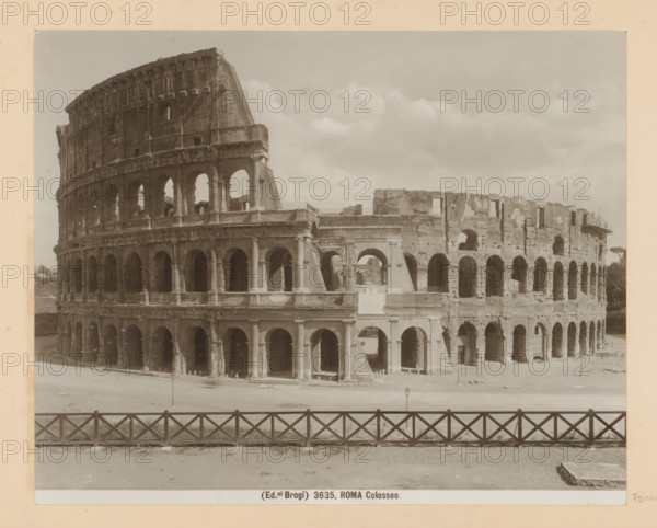 Exterior of the Colosseum in Rome, Roma Colosseo, photograph, Part of Photo album with photographs of landmarks and works of art in Italy., number: '3635', photographer: Edizioni Brogi, Rome, c. 1875 - c. 1907, paper, gelatin silver print, developed gelatin silver print, height 200 mm x width 250 mm, ruin of a building, architecture, Colosseum, A sepia-toned photograph shows the exterior of a large ancient amphitheater with multiple levels of arches and columns, the stone facade weathered and partially ruined, with sections missing and exposed inner walls. The foreground features a low wooden fence with a crisscross pattern running horizontally across the image, and an open paved area between the fence and the building. The sky above is filled with scattered clouds, and small architectural details such as niches, stair openings, and varying arch shapes are visible along the structure.