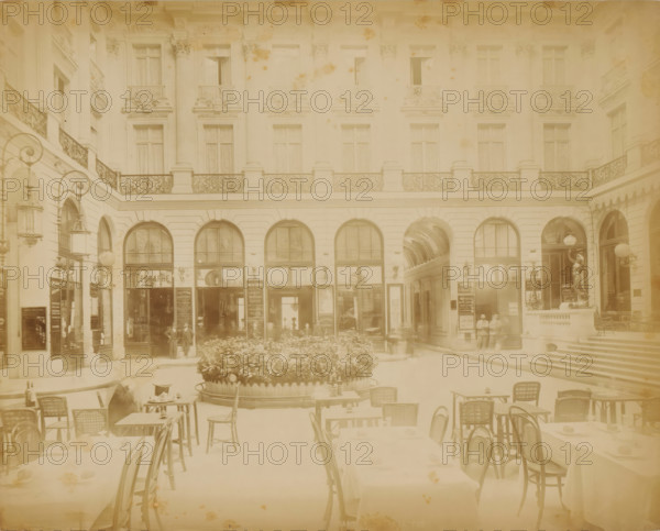 Passage with a terrace in Paris, photograph, Part of Travel Album with photographs of sights in Sweden, Norway, Denmark, Bavaria, Milan and Paris., Paris, c. 1870 - c. 1900, photographer, paper, albumen print, height 221 mm x width 278 mm, pavement, terrace in front of inn or restaurant, advertising, A faded courtyard scene with ornate building facades featuring tall arched doorways and windows, decorative iron railings on a balcony level, sculpted pilasters and ornate cornices, street lamps with curved arms mounted on walls, a covered passage visible through one arch, a raised circular planter filled with dense foliage in the center, café tables and wicker chairs arranged in the foreground with table settings and small objects on them, a few standing figures near the entrances, and a statue on a plinth beside a staircase leading up to another entrance.