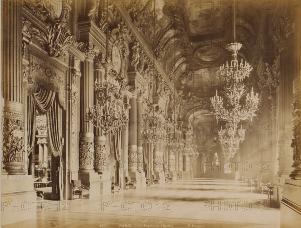 PARIS. - Le Foyer de l'Opéra, Foyer of the Opéra Garnier in Paris, photograph, Part of Travel album with recordings of sights in Sweden, Norway, Denmark, Bavaria, Milan and Paris., number: '149', photographer: X photo., Opéra de Paris, c. 1870 - c. 1890, A long ornate interior space with tall fluted columns topped by elaborate capitals and sculpted reliefs lining one side, heavy draped curtains framing recessed openings, richly carved pilasters and decorative moldings along the walls, multiple large crystal chandeliers suspended from a painted vaulted ceiling with ornate plasterwork and framed ceiling medallions, rows of carved wooden chairs and small tables placed against the walls, intricate floral and foliate carvings on column bases and surrounds, polished patterned wooden flooring reflecting light, and soft shafts of light and shadow stretching across the floor toward a distant arched opening.