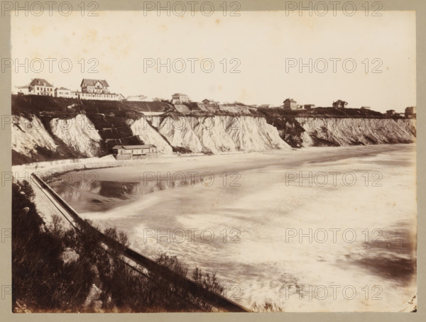 Côte des Basques near Biarritz, album page, photograph, This photo is part of an album., annotation: 'Biarritz, Côte des Basques, ...', check stamp: 'RFK R...', 1880 - 1910, photographer, paper, albumen print, albumendruk, height 174 mm x width 234 mm, rocky coast, civic architecture, edifices, dwellings, Biarritz, A sepia-toned coastal scene showing a wide sandy shoreline with waves washing in and a reflective wet sand area, bordered by steep chalk cliffs with striated white faces and darker vegetation on top, a low seawall curving along the base of the cliffs with a small building next to it, a row of houses and larger multi-story buildings perched along the cliff edge, and foreground shrubbery and a railing descending toward the beach.