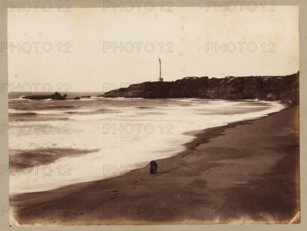 Côte des Fous with lighthouse near Biarritz, album page, photograph, This photo is part of an album., annotation: 'Biarritz, Côte des Fous, ...', 1880 - 1910, photographer, paper, albumen print, height 175 mm x width 234 mm, beach, lighthouse, lightship, Biarritz, A sepia-toned coastal scene showing a sandy beach curving along the shoreline with wet sand reflecting light, gentle waves washing ashore in layered bands, and a rocky headland in the distance topped by a tall, slender lighthouse and a couple of low buildings; a small dark figure stands on the beach near the water's edge, and scattered rocks and a distant rocky outcrop sit offshore beneath a pale sky.