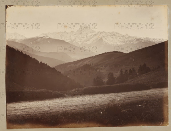 View of Pic du Midi de Bigorre from the Col d'Aspin, album page, photograph, This photo is part of an album., annotation: 'Pic du Midi, vue prise de Col d'Aspin', 1880 - 1910, photographer, paper, albumen print, height 178 mm x width 233 mm, mountains, Midi de Bigorre, Pic du, A sepia-toned landscape photograph showing layered mountain ridges receding into the distance with a snow-capped peak rising centrally in the background, softer distant slopes and valleys between the ridges, darker forested hills in the midground with clusters of conifer trees, a gently sloping foreground plain with textured ground and scattered low vegetation, and a light sky above with subtle blemishes on the photographic surface.