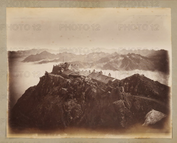 View from Pic du Midi de Bigorre, album page, photograph, This photo is part of an album., annotation: 'Vue prise du Pic du Midi', 1880 - 1910, photographer, paper, albumen print, height 178 mm x width 226 mm, mountains, Midi de Bigorre, Pic du, A sepia-toned photograph showing a rugged rocky mountain peak with a stone building complex and wooden structures perched near the summit, low clouds or mist filling the valleys below, distant jagged mountain ranges visible through the haze, a terrace or platform with railings and flagpoles atop the structure, steep rock faces and boulders in the foreground, and a faded border around the image.