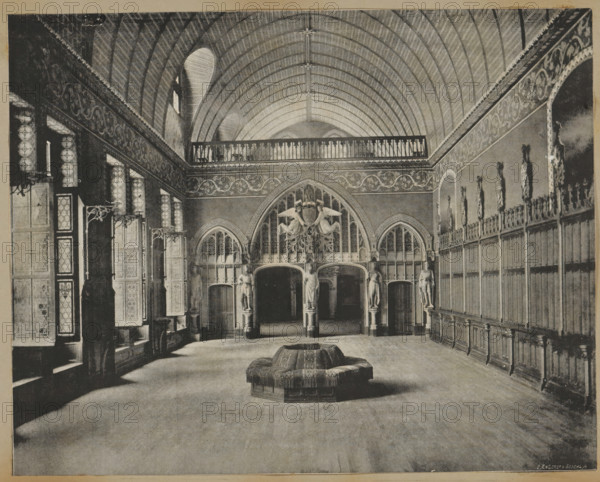 Interior of the castle of Pierrefonds, album page, photomechanical print, This print is part of an album., maker: Angerer & Göschl, C., 1880 - 1900, paper, autotype, height 201 mm x width 248 mm, castle - AA - civic architecture: inside, Pierrefonds, Castle of Pierrefonds, A large vaulted hall with a ribbed arched ceiling and a decorative balcony running along the upper wall is shown, with ornate carved panels and tall narrow windows along one side and a long wooden paneled wall with sculpted figures in niches along the opposite side; at the far end there are arched openings flanked by sculpted statues and an elaborate carved archway and crest, and in the center foreground a circular upholstered seating piece with fringe sits on a broad wooden floor.