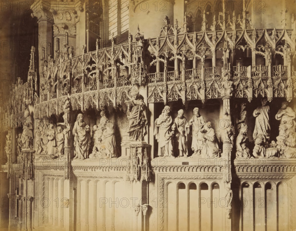 Choir screen of Chartres Cathedral, album page, photograph, This photo is part of an album., annotation: 'Cathédrale de Chartres ...', photographer: Adolphe Giraudon, 1880 - 1910, paper, albumen print, height 215 mm x width 276 mm, rood-screen, choir-screen, iconostasis, Chartres, Chartres Cathedral, A sepia-toned photograph of an ornate carved stone choir screen featuring pointed arches and intricate tracery, with rows of sculpted figures standing on ledges beneath the arches. The upper portion shows a repeating pattern of decorative pinnacles, openwork balustrades, and foliated details, while the lower register presents larger statues of robed figures in varied poses, some holding objects and some grouped together. Fluted vertical panels and arched recesses form the lower wall beneath the sculptures, and slender columns and clustered shafts rise between sections. The lighting casts soft shadows that emphasize the relief carving and the textured surfaces of the stone.