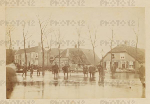 Flooding Nieuwkuyk 1916, Flood in Nieuwkuijk, photograph, photographer: attributed to Albert Greiner, Amsterdam, 1880 - 1881, Attributed to, pasteboard, paper, albumen print, albumendruk, height 100 mm x width 153 mm, height 210 mm x width 256 mm, the year 1916 should be 1880, village, flood, overvloed, Nieuwkuijk, Period, 1880-12-29 - 1880-12-30, A sepia-toned street scene showing a row of houses with thatched and tiled roofs behind a line of leafless trees, a group of people standing and walking on a reflective wet surface in front of the houses, a tall street lamp near the center right, several figures wearing coats and hats, and the buildings' windows and doors visible along the street.