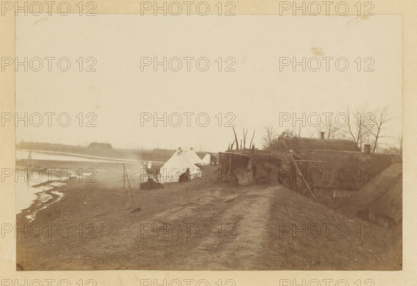 Flood in Nieuwkuijk, Flood Nieuwkuyk N Br. 1916, photograph, title: 'Overstroming Nieuwkuyk N Br. 1916', photographer: attributed to Albert Greiner, Amsterdam, 1880 - 1881, Attributed to, pasteboard, paper, albumen print, albumendruk, height 105 mm x width 155 mm, height 210 mm x width 262 mm, Date 1916 is incorrect, should be 1880, village, flood, overstroming, Nieuwkuijk, Period, 1880-12-29 - 1880-12-30, A sepia-toned scene showing a riverside encampment with several canvas tents pitched along a dirt embankment, a narrow path running between the tents and the camera, and makeshift wooden structures and poles nearby; water and a muddy shoreline appear to the left with a small boat pulled up near the bank, and low buildings and leafless trees are visible in the distant background on the right.