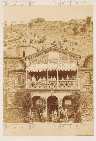Entrance to the Grotta Giusti in Pistoia, photograph, photographer: Scevola Ghelli, Pistoia, 1860 - 1900, Tuscany, pasteboard, paper, albumen print, albumendruk, height 201 mm x width 139 mm, height 250 mm x width 224 mm, watering-place, spa, Grotta Giusti, A sepia-toned photograph showing a stone building with a balcony and an awning, the balcony edge decorated with a pattern of openings and supported by posts, a group of people standing and sitting along the balcony railing, and additional people posed on the ground level in front of arched doorways framed by foliage; the building has shuttered windows, a small round tower rising to one side with people visible on its top, and a hillside covered in sparse vegetation visible behind the structure.