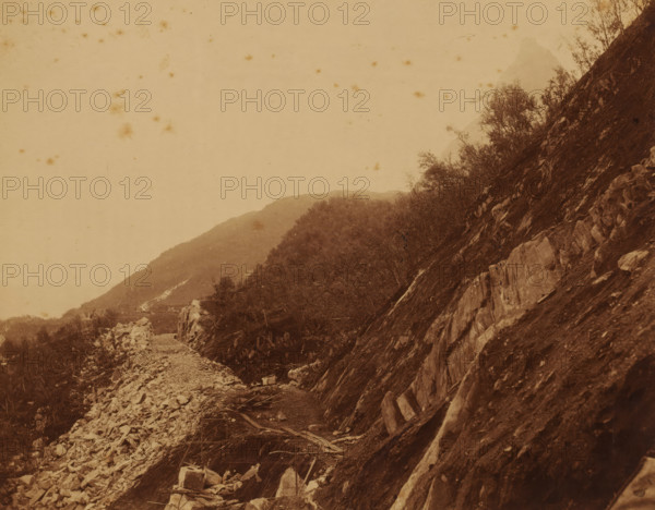 Paved mountain path for the construction of the Luleå - Narvik railway line, photograph, Sweden, 1888 - 1900, photographer, pasteboard, paper, albumen print, height 196 mm x width 249 mm, height 200 mm x width 259 mm, road, path, A sepia-toned landscape photograph showing a rocky hillside sloping steeply on the right with exposed rock faces and scattered vegetation, a rough gravel path or cleared area running along the base of the slope with loose stones and wooden planks, shrubs and small trees growing on the lower slopes and along the midground, distant rounded hills fading into the background, and a pale sky with small dark specks and stains across the image.