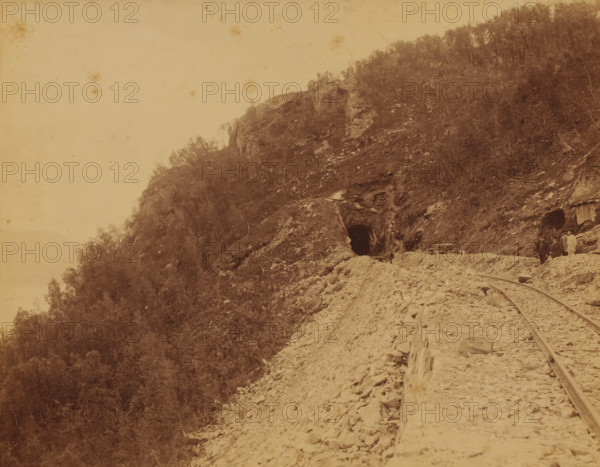 View of the Luleå - Narvik railway line, on the right some railway workers, photograph, Sweden, 1888 - 1900, photographer, pasteboard, paper, albumen print, height 195 mm x width 253 mm, height 204 mm x width 259 mm, railway, train, Nordland, Luleå, Narvik, A sepia-toned landscape showing a rocky hillside with sparse trees and vegetation, a dark tunnel opening carved into the rock near the center, rough gravel and debris leading toward the tunnel, narrow-gauge rails running along the ground toward the right side, a small group of people standing near the tunnel entrance and beside another darker opening with a wooden structure, scattered rocks and wooden planks along the path, and an open pale sky occupying the upper left portion of the image.