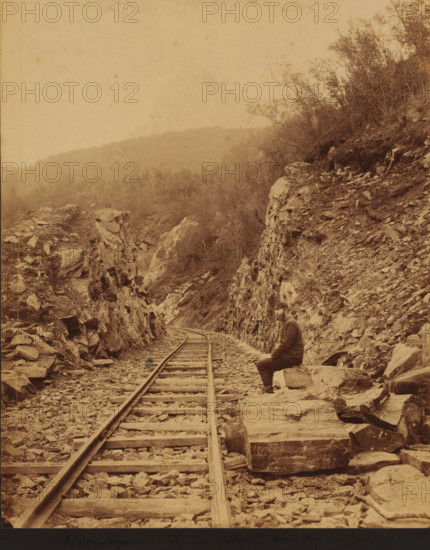 View of the Luleå - Narvik railway line, on the right an unknown man along the track, Norway, Lulea - Ofoten railway, photograph, Sweden, 1888 - 1900, photographer, pasteboard, paper, albumen print, height 224 mm x width 197 mm, height 262 mm x width 205 mm, railway, train, Nordland, Luleå, Narvik, A sepia-toned photograph showing a narrow railway track running into a rocky gorge with jagged rock faces rising on both sides, loose stones and wooden sleepers scattered along the track, a man wearing a coat and hat seated on a large rock near the right rock face looking toward the camera, sparse vegetation and small trees clinging to the slopes, distant tree-covered hills visible through a hazy sky.