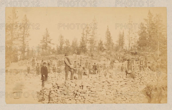 Group of men, possibly miners in Northern Sweden between Luleå and Narvik, photograph, Sweden, 1891 - 1900, photographer, pasteboard, paper, albumen print, height 121 mm x width 203 mm, height 240 mm x width 333 mm, miner, A wide sepia photograph shows a rocky outdoor scene with a group of people standing among piles of irregular stones and low stacked stone walls, tall evergreen trees rising in the background, several men wearing hats and work clothes scattered across the rocky ground, a few individuals standing on higher stone piles, one man in the foreground facing the camera holding an object, another man nearby standing with a tool, and additional figures dispersed along terraces of stone with trees and rough terrain beyond.