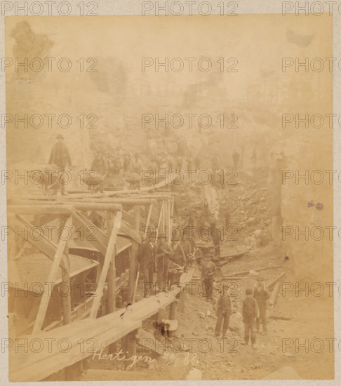 Works in the Hertigen mine near Malmberget, Sweden, photograph, Malmberget, 1888-08-20, photographer, pasteboard, paper, albumen print, albumendruk, height 182 mm x width 164 mm, height 332 mm x width 242 mm, quarry (stone), Hertigen, A faded sepia photograph showing a group of men standing and walking on a rough, rocky worksite with wooden scaffolding and planks forming a raised walkway along one side; some men stand on the walkway while others are on the uneven ground below, many wearing hats and work clothes, and a few appear to be dressed more formally; large wooden beams and structural supports are visible in the foreground, with piles of rocks and timbers scattered throughout the scene and a steep rocky face rising at the back where additional figures stand on ledges.