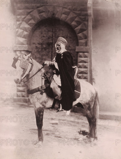 Officer on horseback, probably in Algiers, Officer Indischene, photomechanical print, inscription: 'foto Leroux Alger', photographer: Alexandre Leroux, Algiers, cliché maker: anonymous, 1880 - 1900, North Africa, pasteboard, paper, collotype, photoprint, height 317 mm x width 247 mm, riding a horse, ass, or mule, rider, horseman, A sepia-toned photograph shows a man seated on a horse in front of a large stone archway with a textured pattern. The man wears a wrapped head covering and a long dark cloak draped over lighter garments, and he has a visible beard. The horse is outfitted with ornate decorative tack including a patterned headpiece and a decorated breast collar, and the saddle area shows embroidered fabric and straps. The ground beneath the horse appears uneven and there are faint shadows and marks on the stone surface and surrounding wall.
